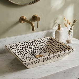 May include: A rectangular vessel sink featuring a cream-coloured base with a repeating pattern of irregular black spots. The sink includes a central gold-toned drain fixture and is positioned on a white marble countertop with a wall-mounted gold tap.