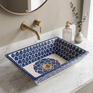 May include: A rectangular ceramic vessel sink featuring a hand-painted blue and white fish scale pattern on the interior walls and a central floral motif at the base. The sink includes a gold-toned metal drain and is set on a white marble countertop.