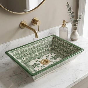 Puede incluir: Un lavabo rectangular de cerámica con base blanca, decorado con complejos patrones geométricos verdes y un motivo floral central. El lavabo incluye un desagüe de metal dorado y se combina con un grifo de pared a juego en tono dorado.