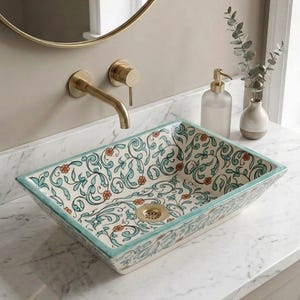 May include: A rectangular ceramic vessel sink featuring a hand-painted pattern of teal scrolling vines and small orange flowers on a white background. The sink includes a gold-toned drain and is paired with a matching gold-toned wall-mounted faucet.