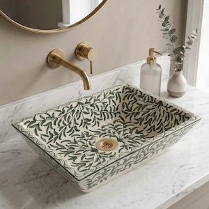 May include: A rectangular vessel sink featuring a hand-painted dark green botanical leaf pattern on a cream-colored base. The sink includes a gold-toned drain and is paired with a wall-mounted gold faucet, a frosted glass soap dispenser, and a small vase.