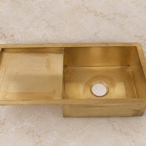 Op de afbeelding: Een rechthoekige gootsteen van gepolijst messing met een metallic gouden afwerking. De gootsteen is voorzien van een enkele bak aan de rechterkant en een geïntegreerd plat afdruiprek aan de linkerkant, met een ronde afvoeropening in het midden.