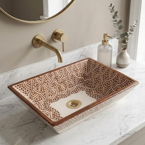 May include: A rectangular vessel sink featuring a brown and white geometric pattern on the interior and exterior walls. The sink has a smooth, off-white base at the bottom with a circular gold-toned drain cover, set against a white marble countertop.