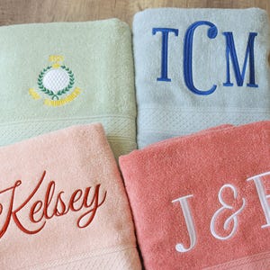 May include: Four personalised bath towels in pastel shades. One is pale green with a golf emblem, another is light blue with the initials TCM, a third is pink with the name Kelsey, and the last is coral with the initials J&B.