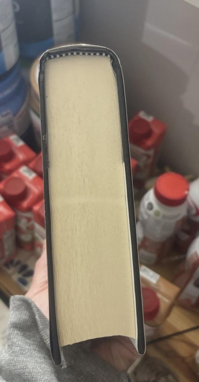 May include: A hardcover book with a light beige page block and a black spine. The pages are visible from the side, showing a textured surface. The book's edges are rounded.