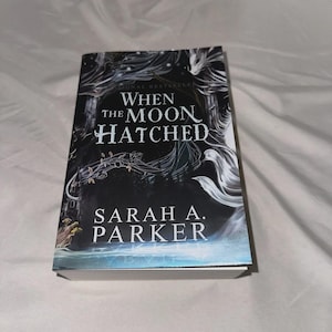 May include: A hardcover book titled "When the Moon Hatched" by Sarah A. Parker. The cover features a dark, fantastical design with the title in white and the author's name in a clean font. The book is set against a white background.