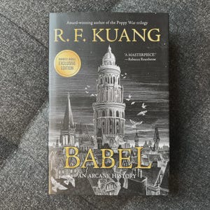 May include: A hardcover book titled "BABEL: An Arcane History" by R.F. Kuang. The cover features a detailed illustration of a tall tower and cityscape in gold and black. Includes a Barnes & Noble Exclusive Edition sticker.