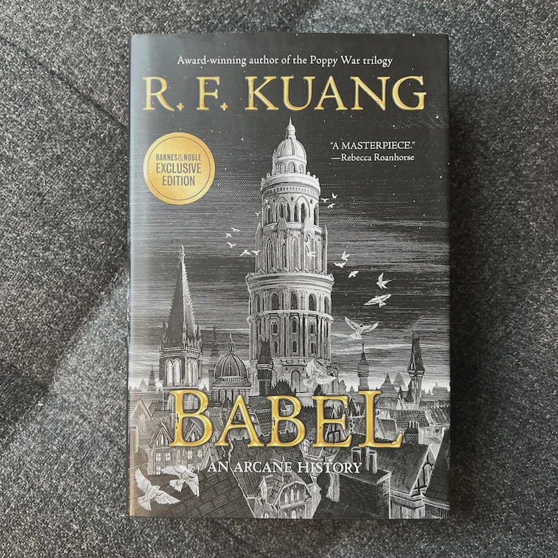 May include: A hardcover book titled "BABEL: An Arcane History" by R.F. Kuang. The cover features a detailed illustration of a tall tower and cityscape in gold and black. Includes a Barnes & Noble Exclusive Edition sticker.