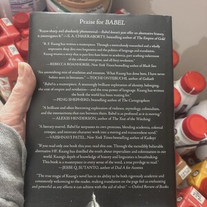 May include: The back cover of the book "Babel" is shown, featuring quotes praising the novel. The cover is black with white text, including the title and author's name. The book's ISBN is visible at the bottom. The book is held by a person.