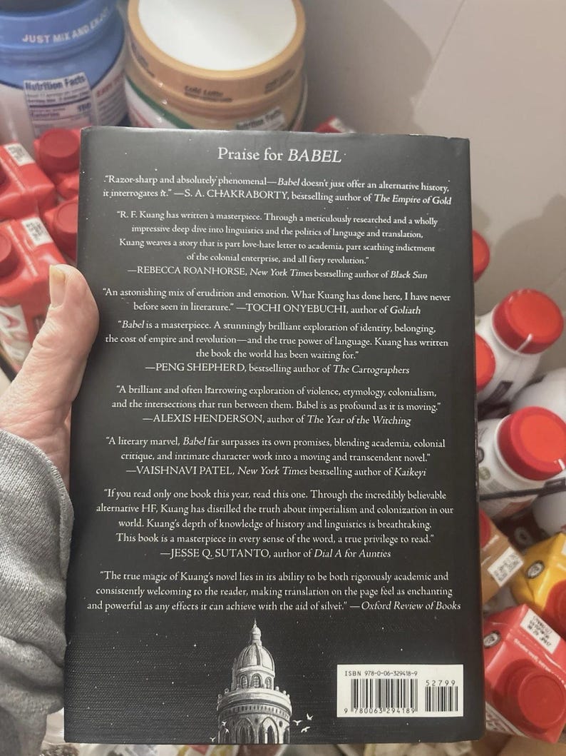 May include: The back cover of the book "Babel" is shown, featuring quotes praising the novel. The cover is black with white text, including the title and author's name. The book's ISBN is visible at the bottom. The book is held by a person.