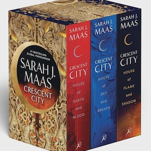 May include: A boxed set of three books by Sarah J. Maas, titled "Crescent City." The book spines are red, blue, and orange, with the author's name and series title in white. The box has a gold and navy blue ornate design.