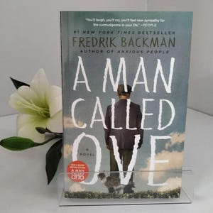 May include: A paperback book titled A Man Called Ove by Fredrik Backman. The cover features a man in a dark suit standing in a field under a cloudy sky, with a small cat near his feet. Text includes #1 New York Times Bestseller and A Major Motion Picture.
