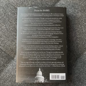 May include: The back cover of a book titled "BABEL" with a dark background. The cover features quotes praising the book from various authors and publications. The book's ISBN and a barcode are visible at the bottom.