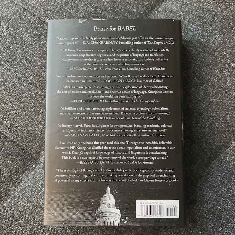 May include: The back cover of a book titled "BABEL" with a dark background. The cover features quotes praising the book from various authors and publications. The book's ISBN and a barcode are visible at the bottom.