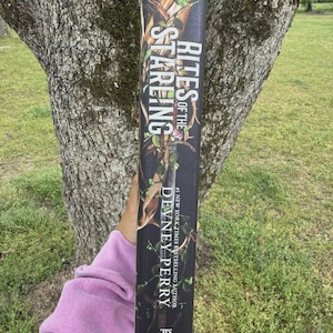 May include: A book titled "Rites of the Stareng" by Devney Perry. The spine features a dark background with green foliage and the author's name in white. The book is held in front of a tree.