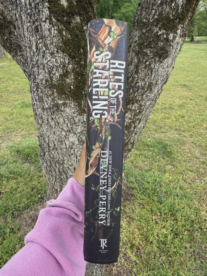 May include: A book titled "Rites of the Stareng" by Devney Perry. The spine features a dark background with green foliage and the author's name in white. The book is held in front of a tree.