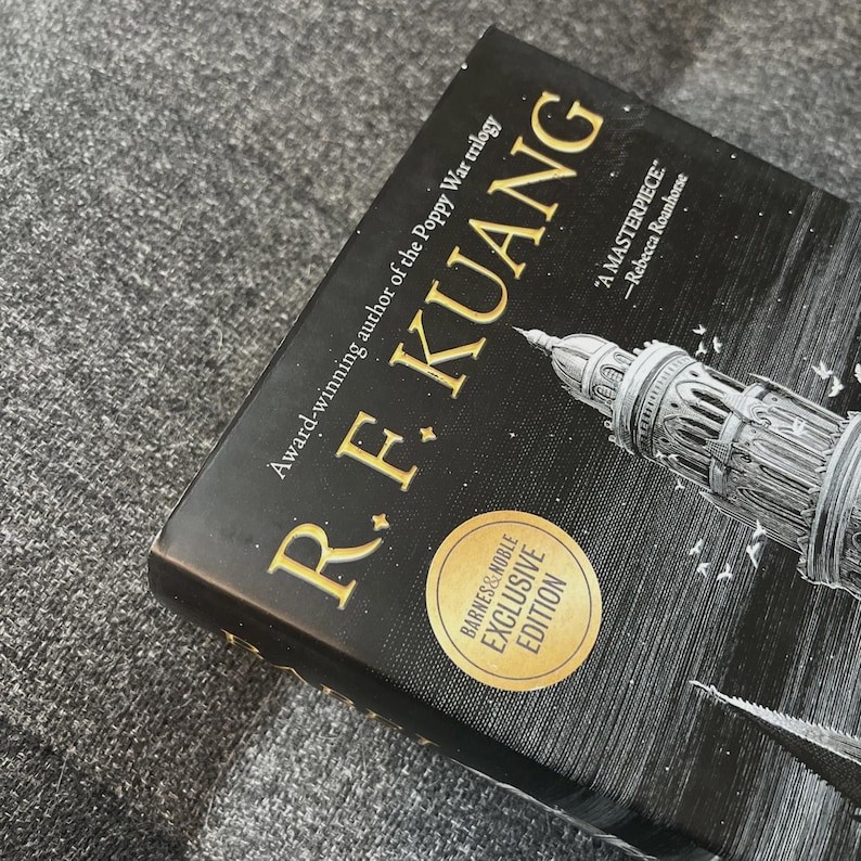 May include: A hardcover book by R.F. Kuang, with gold lettering and a black cover. The book is a Barnes & Noble Exclusive Edition. The cover features a detailed illustration of a building and the quote "A MASTERPIECE" by Rebecca Roanhorse.