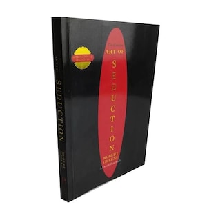 May include: A black book titled The Art of Seduction by Robert Greene with a large red vertical oval on the cover. The text is printed in gold and white. A gold circular sticker on the upper left corner reads International Bestseller.