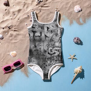 May include: A one-piece swimsuit featuring a black and white graffiti-style print with abstract lettering and drips. The garment has a scoop neckline and white trim along the armholes and neckline, providing a clean contrast to the dark pattern.