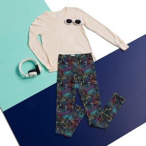 May include: A long-sleeve cream-colored shirt, black leggings with a multicolored circuit board pattern, white oval sunglasses, and white over-ear headphones. The leggings feature a dark background with vibrant blue, pink, and yellow lines.