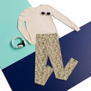 May include: A cream-colored long-sleeve shirt, a pair of beige leggings with a repeating black line-art pattern of various vintage-style illustrations, a pair of white oval-framed sunglasses, and white over-ear headphones with black accents.