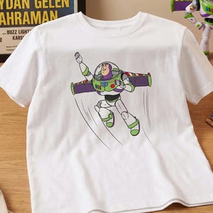 Puede incluir: Camiseta blanca con un gr&aacute;fico de Buzz Lightyear en vuelo, con detalles verdes y morados. La camiseta est&aacute; sobre una superficie de madera, junto a figuras de Toy Story, c&oacute;mics y pegatinas del logo de Pixar. Un p&oacute;ster turco es visible.