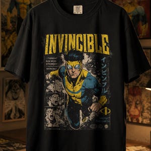 Op de afbeelding: Een zwart t-shirt met korte mouwen en een grafische print van het personage Invincible. Het ontwerp bevat de tekst INVINCIBLE, I THOUGHT YOU WERE STRONGER en PROTECT THE EARTH, samen met stripboekpanelen en Japanse karakters.