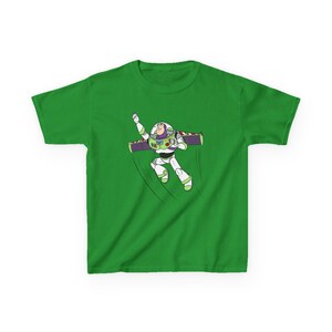 Puede incluir: Una camiseta verde con una ilustraci&oacute;n de dibujos animados de Buzz Lightyear en una pose de vuelo. El personaje lleva un traje espacial blanco y verde con alas moradas. El dise&ntilde;o est&aacute; sobre un fondo verde liso.
