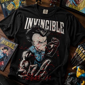 May include: A black short-sleeve t-shirt featuring a graphic print of the character Invincible with a spiky hairstyle, blue and black suit, and red blood splatters. The word INVINCIBLE is printed in large white capital letters above the character.