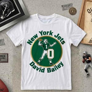 Op de afbeelding: Wit T-shirt met een groen en goud New York Jets logo met een American football speler en de naam "David Bailey". Ook een ingelijste Raiders helm, een voetbal en andere voetbal-thema memorabilia.