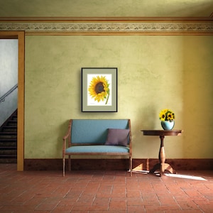 May include: A framed print of a sunflower with yellow petals and a brown center. The print is hanging on a green wall above a blue couch and a wooden table with a vase of yellow flowers.