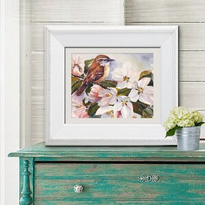 May include: A framed watercolor painting of a small brown bird perched on a flowering branch with white and pink blossoms. The white frame has a textured border. The painting is displayed on a teal-colored wooden cabinet.