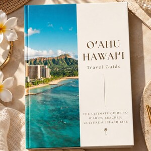 May include: A hardcover travel guide book titled O'AHU HAWAI'I. The cover features a vertical photograph of a coastal landscape with a mountain, high-rise buildings, and turquoise ocean water. The text reads The Ultimate Guide to O'ahu's Beaches, Culture & Island Life.