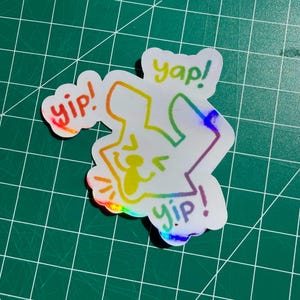 May include: A holographic sticker featuring a cartoon character with rainbow outline and the words "yip!" and "yap!". The sticker has a shiny, iridescent finish, and is placed on a green cutting mat.