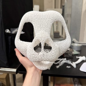 May include: A white, open-faced mask with a textured, web-like design. The mask has large eye openings and a prominent nose area. The object is held in a person's hand, with a blurred background of a workshop.