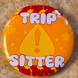 May include: A round orange button with the words "TRIP SITTER" in white bubble letters. The button has a yellow triangle with an exclamation point and a leaf. The button has red and yellow stripes and yellow stars.