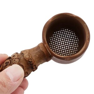 Könnte beinhalten: Ein braunes Teesieb aus Holz mit einem zylindrischen Kopf, einem perforierten Metallsieb als Boden und einem strukturierten, knorrigen Griff im Bambus-Stil. Die natürliche Holzmaserung ist durchgehend sichtbar und betont die organische Textur.