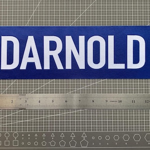 May include: A rectangular blue sticker with the white text DARNOLD in a bold sans-serif font. A metal ruler is positioned directly below the sticker, showing a length of approximately 13 inches for the text label.