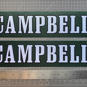 May include: Two rectangular dark green decals featuring the word CAMPBELL in bold white capital letters with a black outline. A metal ruler is positioned below the decals, showing a length of approximately 16 inches for the text strips.