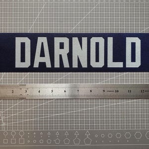 May include: A rectangular dark blue sticker featuring the white text DARNOLD in a bold, sans-serif font. A stainless steel ruler is positioned directly below the sticker, showing a length of approximately 38 centimetres for the decal.