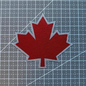 May include: A red maple leaf patch with a textured surface and a light grey border. The patch is shaped like a classic maple leaf with five distinct points and a small stem at the bottom, featuring a consistent woven fabric appearance throughout.