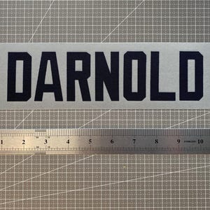May include: A rectangular white decal featuring the word DARNOLD in bold, dark navy blue block lettering. A stainless steel ruler is positioned directly below the decal, showing the text spans approximately 10.5 inches in total length.