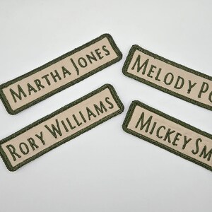 May include: Four rectangular fabric name patches with a beige background and olive green embroidered borders and lettering. The patches feature the names Martha Jones, Melody Pond, Rory Williams, and Mickey Smith in a clean, sans-serif font.