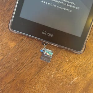 May include: A Kindle e-reader with a clear protective case rests on a wooden surface. The screen displays text: "Ten suspects. One locked bank. Everyone is a thief, but who is a murderer?" A small charm with a book design hangs from the device.