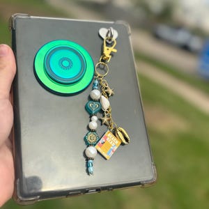 Puede incluir: Una funda transparente para teléfono con un soporte para teléfono verde y azul y un llavero dorado. El llavero tiene cuentas azules, blancas y doradas, un dije de concha y un pequeño dije de libro que dice "Beach Read".
