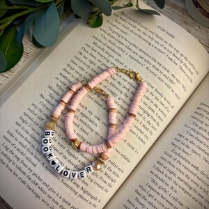 May include: A bracelet made of pink and tan clay heishi beads with gold-toned accents and white letter beads spelling out BOOK LOVER with a black heart symbol. The bracelet features a gold-toned lobster claw clasp and is resting on an open book.