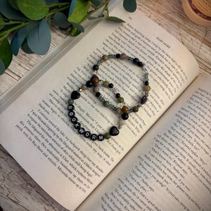 May include: Two beaded bracelets resting on an open book page. One bracelet features black, green, and brown beads with a heart-shaped charm. The other bracelet includes white letter beads spelling BOOKWORM, black heart beads, and green beads.