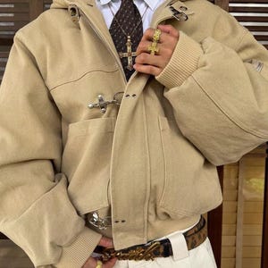 Puede incluir: Chaqueta con capucha beige con cierre de palanca, usada sobre una camisa blanca y una corbata estampada. El atuendo incluye un colgante de cruz dorada, anillos dorados y un cinturón estampado con hebilla dorada. La chaqueta tiene puños acanalados.
