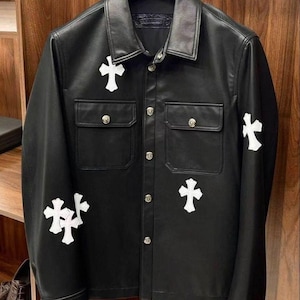 Puede incluir: Una chaqueta de cuero negro con cuello clásico, mangas largas y cierre frontal de botones. La prenda está adornada con varios parches blancos en forma de cruz en el pecho, las mangas y los paneles frontales, con botones plateados.