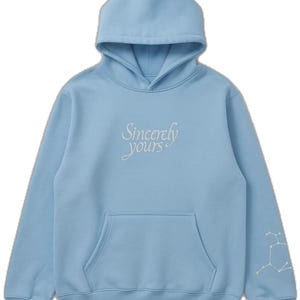 Op de afbeelding: Een lichtblauwe hoodie met een voorzak en een capuchon. De tekst "Sincerely yours" is op de borst geborduurd. Een sterrenbeeldontwerp is op de linkermouw geborduurd. De hoodie is gemaakt van zacht materiaal.